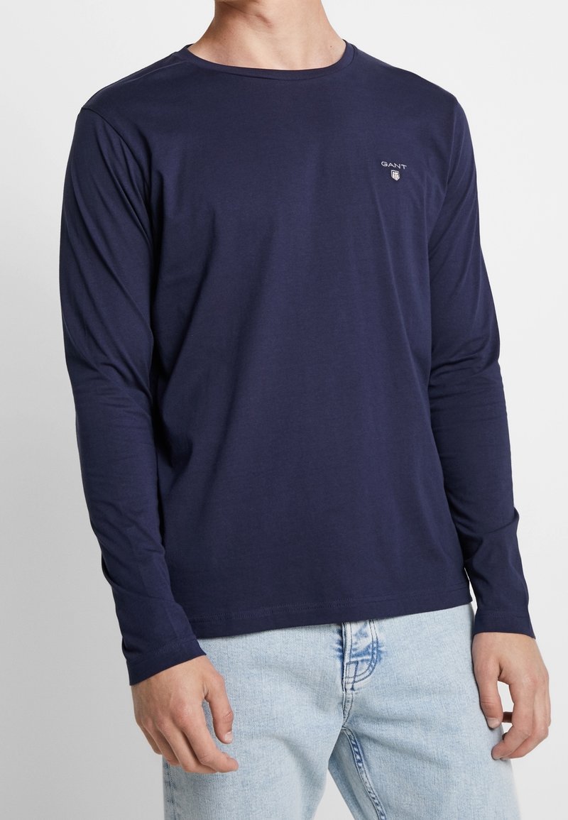 Marineblaues Langarmhemd aus glattem Stoff, mit Rundhalsausschnitt und einem kleinen gestickten Logo auf der Brust. Kombiniert mit hellblauen Jeans.