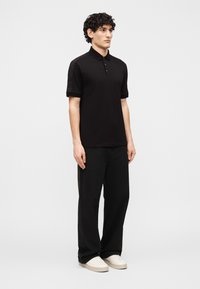 Νεαρός άνδρας όρθιος, φοράει μαύρο πόλο μπλουζάκι με κοντά μανίκια, μαύρο άνετο παντελόνι και λευκά αθλητικά παπούτσια, μπροστά σε απλό φωτεινό φόντο.