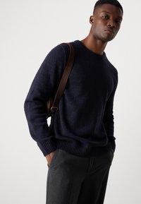 Navy knit sweater with textured surface, round neck, and ribbed hem, paired with gray trousers and a brown shoulder strap bag.