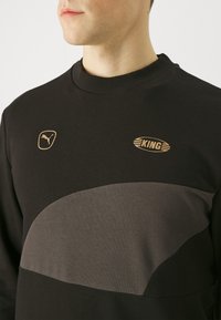 Felpa nera con un pannello curvo grigio scuro. Presenta loghi dorati: Puma a sinistra e 'KING' con linee sul petto. Collo a costine.
