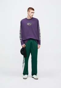 Violeta krekls ar garām piedurknēm, centrā ar logo un uzdrukātiem grafiskiem elementiem uz piedurknēm, kopā ar zaļām sporta biksēm ar sānu svītrām un baltiem akcentiem.