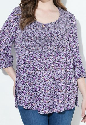 Woman wearing a purple floral print blouse with 3/4 sleeves and smocked detail at the bust, paired with blue jeans.