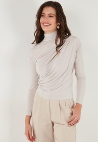 Licht beige lange mouwen top met een hoge hals en gedrapeerde textuur. Kenmerkt zich door zachte, rekbare stof met geplooide details aan één kant.
