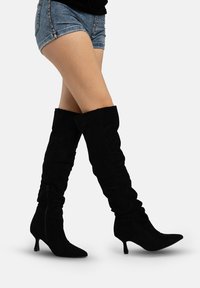 Zwarte suède laarzen over de knie met een nonchalant ontwerp en een puntige neus, voorzien van een kleine hak en een ritssluiting aan de zijkant.