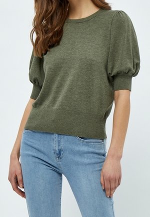 Kvinde iført olivengrøn sweater med pufærmer kombineret med lyseblå jeans, stående mod en ensfarvet lys baggrund.