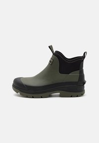 Zöld gumicsizma fekete felsőrésszel, texturált díszítéssel és robusztus fekete talppal, amely mély barázdákkal rendelkezik a tapadás érdekében. Barbour logó az oldalon.