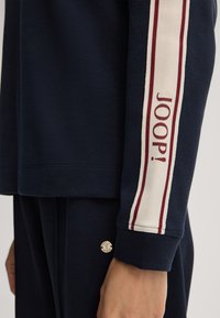 Marineblauwe sweatshirt met een witte en bordeauxrode streep langs de mouw en de tekst "JOOP!". Zacht materiaal met een relaxte pasvorm en ribgebreide manchetten.