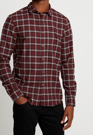 Mann trägt ein bordeauxrotes und graues kariertes Hemd mit Knöpfen und schwarze Hose, steht mit einer Hand in der Tasche vor weißem Hintergrund.
