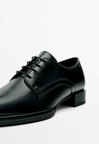 Musta kiillotettu nahkakenkä, jossa on suljettu nauhoitus ja matala korko, yksinkertaisella valkoisella taustalla.