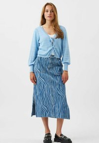 Világoskék kötött kardigán, elöl megkötős, hosszú farmer szoknyával párosítva, amely világosabb kék árnyalatú zebra mintával díszített. Fekete slip-on cipők.