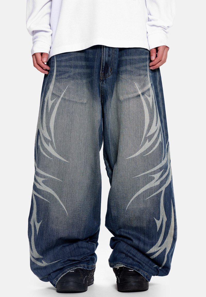 Φαρδιά τζιν σε σκούρο μπλε χρώμα με γκρι φυλετικά σχέδια στα πόδια. Εφαρμοστή γραμμή στον αστράγαλο. Φοριέται με μαύρα αθλητικά παπούτσια.