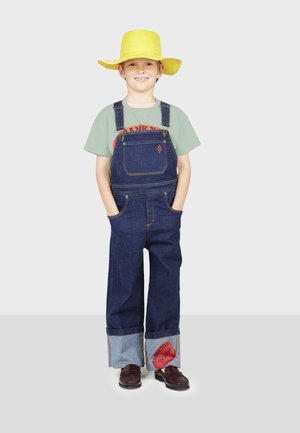 Denim kopējie ar brīvu piegriezumu, zila krāsa un regulējamas siksnas, saskaņoti ar gaiši zaļu krekli un plašu dzeltenu cepuri. Sarkanā akcenta uzlīme.