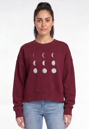 Femme aux cheveux foncés attachés en queue de cheval, portant un sweat bordeaux avec des motifs des phases de la lune et un jean bleu clair, debout devant un fond blanc.