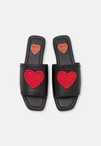 Black slide sandals with a flat sole, featuring a large red crocheted heart on each strap and a gold heart logo on the insole.