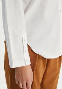 Camisa blanca de manga larga con botones en la muñeca de una persona que lleva pantalones marrones, con la mano relajada a su lado.
