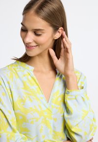 Leichte Bluse in Pastellblau mit einem lebhaften gelben Blumenmuster. Verfügt über einen V-Ausschnitt und lange, gepuffte Ärmel mit Knopfakzenten.