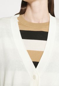 Bílý kardigan s výstřihem do V a zapínáním na knoflíky, nošený přes pruhovaný pletený top s hnědými, černými a béžovými vodorovnými pruhy.