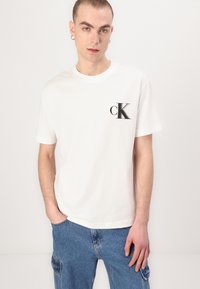White cotton t-shirt with a round neckline, short sleeves, and a black "cK" logo on the left chest. Worn with blue cargo jeans.