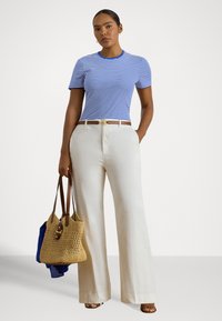 Γυναίκα που φοράει μπλε και λευκό ριγέ κοντομάνικο μπλουζάκι, μπεζ παντελόνι με φαρδιά μπατζάκια, καφέ ζώνη, σανδάλια, κρατώντας μια πλεκτή τσάντα tote με ένα μπλε ύφασμα μέσα.