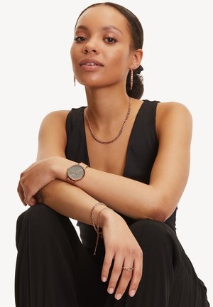 Montre en or rose avec un cadran sombre ; collier chaîne assorti, bracelet et boucles d'oreilles ; textures lisses et motifs entrelacés. Haut noir.