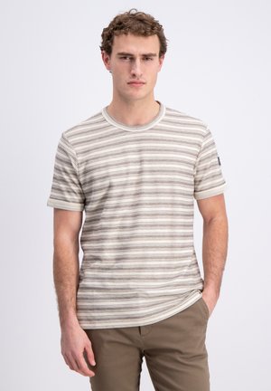 Jonge man met krullend haar, die een beige-wit gestreept T-shirt met korte mouwen en een bruine broek draagt, staand tegen een effen lichte achtergrond.