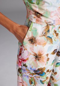 Floraler Jumpsuit mit pastellfarbenen Blüten auf einem weißen Hintergrund, mit taillierter Passform, großen Seitentaschen und glattem Stoff.