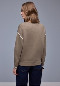 Femme aux cheveux châtain clair mi-longs portant un pull taupe avec des rayures blanches sur les manches et un jean bleu foncé, vue de profil.