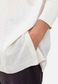 Pull léger en maille de couleur crème avec bord-côte aux ourlets et aux poignets, doté d'une poche latérale et d'une coupe décontractée, présenté sur un tissu foncé.