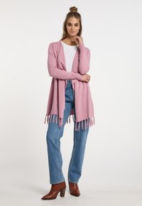 Cardigan rosa chiaro con orlo frangiato, apertura frontale e polsini a coste, indossato sopra una camicia bianca e abbinato a jeans blu a vita alta e stivali marroni.