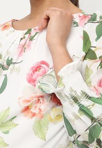 Blouse à fleurs sur fond blanc, ornée de motifs floraux roses et verts. Manches smockées avec poignets et encolure volantés.