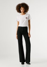 Fehér pamut póló, színes ajak grafikával, magas derekú, fekete, széles lábú farmernadrággal, amelynek oldalán gombos részletek találhatók.