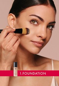 Foundation-Flasche mit schwarzem Deckel auf einem roten Banner. Eine Person trägt flüssige Foundation mit einem Pinsel auf und zeigt dabei glatte Haut und natürliches Make-up.