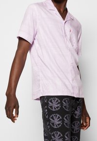 Man wearing a light purple short-sleeve button-up shirt with subtle patterns and black pants with circular graphic prints.