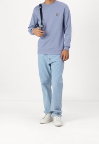 Ljuslila sweatshirt med en gul fjärilslogotyp, parat med ljusblå jeans och vita sneakers, mot en enkel bakgrund.