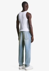 Man in een wit tanktop, lichtblauwe losse jeans, witte sneakers en een zilveren ketting, staand met zijn rug naar de camera tegen een witte achtergrond.