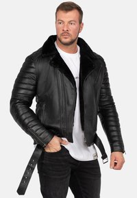 Zwarte leren jas met een nepbont kraag, gequiltte schouderdetails, twee zijritsen en een ceintuur om de taille. Draagbaar over een wit shirt.