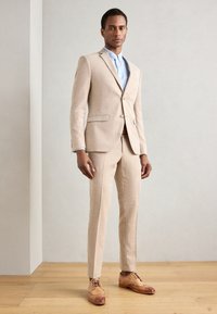 Completo beige con vestibilità slim, tessuto texturizzato, rever a scollo a V e due bottoni. Camicia azzurra chiara sotto, abbinata a scarpe brogue marroni.