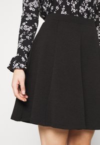 Schwarzer strukturierter Rock mit Falten, aus einem gewebten Material gefertigt. Kombiniert mit einer gemusterten Bluse mit einem blumenförmigen Muster in Schwarz und Weiß.