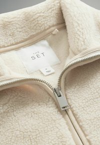 Krämbelagd fleecejacka med en silverdragkedja och slät innerfoder. Etiketten visar "THE SET" i storlek "M." Mjuk, texturerad tyg.