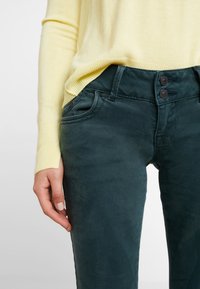 Mujer vestida con pantalones verde oscuro con cierre de doble botón y un suéter de manga larga amarillo pálido, con la mano descansando a un lado.