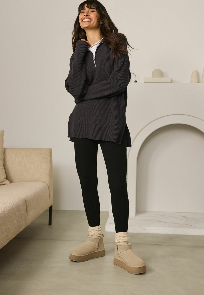 Oversized ciemnoszary sweter dzianinowy z zamkiem przy szyi, czarne legginsy oraz beżowe botki za kostkę na zamek z platformą. Gładkie, teksturowane materiały.