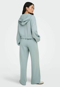 Frau, die seitlich steht, trägt hellblaue Schlaghosen und eine passende kurz geschnittene Kapuzenjacke mit herabgelassener Kapuze, weiße Sneaker, vor neutralem Hintergrund.