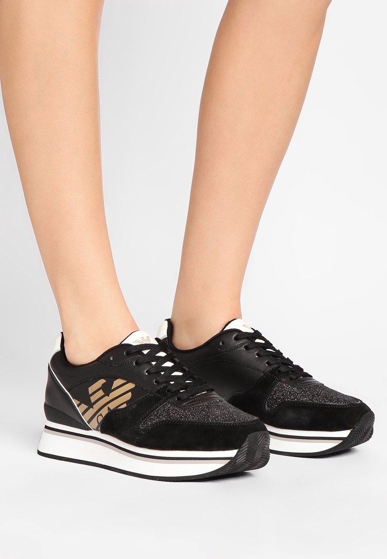 Baskets noires avec semelles intermédiaires blanches et logo doré portées sur des jambes nues sur un fond blanc.