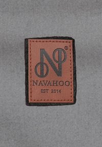 Brun lædermærke med præget logo, "NAVAHOO," og "EST 2014." Omgivet af en sort filt kant. Tydelig syningsdetalje.