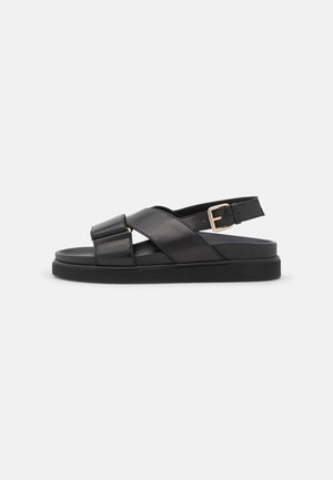 Schwarze Leder-Sandalen mit gekreuztem Design, verstellbarem Knöchelriemen, flacher Sohle und goldfarbener Schnallenverzierung. Glatte Textur, minimalistischer Stil.