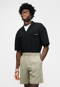 Μαύρο κοντομάνικο πουκάμισο με υφή και δύο τσέπες, σε συνδυασμό με σορτς με ελαστική μέση σε ανοιχτό πράσινο χρώμα. Διαθέτει μικρό λογότυπο στο πουκάμισο.