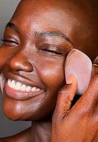 En sminke-svamp i en hånd blir brukt for å jevne ut huden på et smilende ansikt. Svampen er rosa med en rund form og myk tekstur.