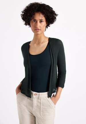 Cardigan verde scuro sopra una maglietta blu navy, caratterizzato da una scollatura drappeggiata e maniche a tre quarti, abbinato a pantaloni beige chiaro. Texture morbida.