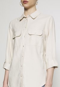 Camisa abotonada de color beige claro con cuello, dos bolsillos en el pecho y mangas remangadas. Elaborada con un tejido suave y de textura lisa.