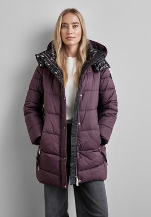 Lila pufferjacka med huva i svart quiltat mönster, sidofickor med dragkedja och tryckknappar. Tyget är mjukt, strukturerat och isolerat för värme.